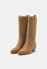 Bottes de cowboy en suède tan avec des détails brodés, bout pointu et talon empilé. Comprend des poignées pour un enfilage facile.