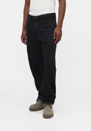 Personne portant un jean noir ample, un haut blanc sans manches et des bottes à lacets marron clair, se tenant devant un fond blanc uni.