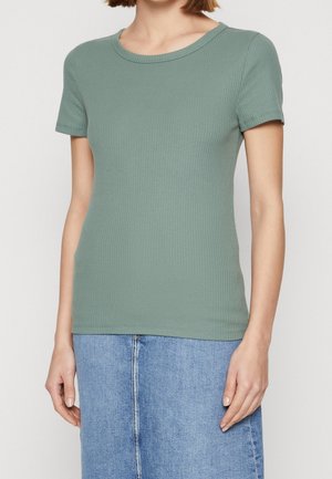 Vrouw met een aansluitend, ribgebreid, saliegroen topje met korte mouwen, gecombineerd met een lichtblauwe, hooggesneden denimrok.