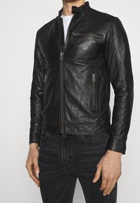 Man med en åtsittande svart läderjacka med tre synliga dragkedjefickor, som är kombinerad med svarta jeans och en vit skjorta under.