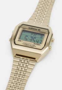 Montre numérique dorée avec un cadran carré, un bracelet en maille métallique et un écran LCD affichant l'heure, la date et le logo de la marque. Comprend des boutons lumière et mode.