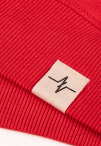 Gorro de punto acanalado rojo con una etiqueta de tela crema que presenta un diseño de línea negra. La textura es suave y elástica, diseñado para mantener el calor.