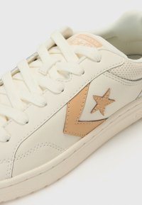 Cream-colored sneaker made of leather with a textured finish, featuring a tan star accent and off-white laces, with a low-profile design.