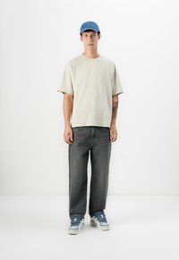 Lys beige oversized t-shirt, straight-leg mørkegrå jeans og blå kasket. Hvide sneakers med blå detaljer, stående mod en ensfarvet baggrund.