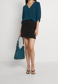Maglietta azzurro chiaro a maniche lunghe con scollatura a V, abbinata a una minigonna nera in suede. La modella tiene in mano una borsa azzurro chiaro con dettaglio in catena dorata e indossa sandali neri.