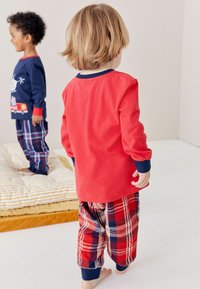 Chemise rouge à manches longues avec des accents navy, associée à un pantalon de pyjama à carreaux rouge et bleu ; des enfants se tiennent sur une surface blanche près d'un coussin rayé.