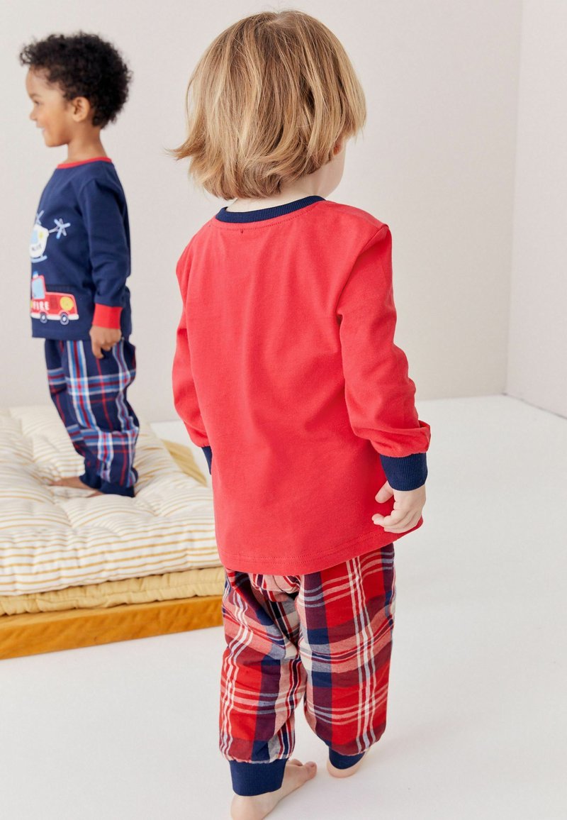 Chemise rouge à manches longues avec des accents navy, associée à un pantalon de pyjama à carreaux rouge et bleu ; des enfants se tiennent sur une surface blanche près d'un coussin rayé.