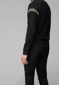 Maglione nero a girocollo realizzato in tessuto testurizzato, con tre strisce orizzontali beige sulla parte superiore della schiena. Abbinato a pantaloni neri.