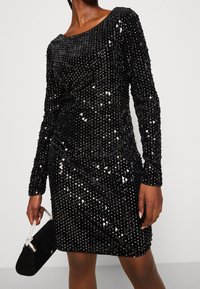 Czarna sukienka z cekinami, z długimi rękawami, w fasonie kopertowym, o dopasowanym kroju, z miękką fakturą i błyszczącymi zdobieniami.