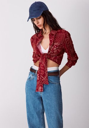 Kobieta w niebieskiej czapce, czerwonej koszuli z wzorem paisley, białej bluzce pod spodem i niebieskich, wysokich dżinsach z brązowym paskiem, patrząca w dół.