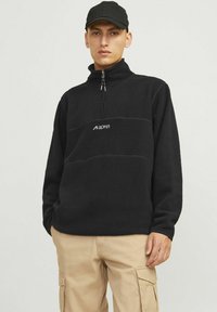 Black fleece quarter-zip pullover features a soft texture, contrasting horizontal seam, and white logo on the chest. Worn with beige cargo pants.