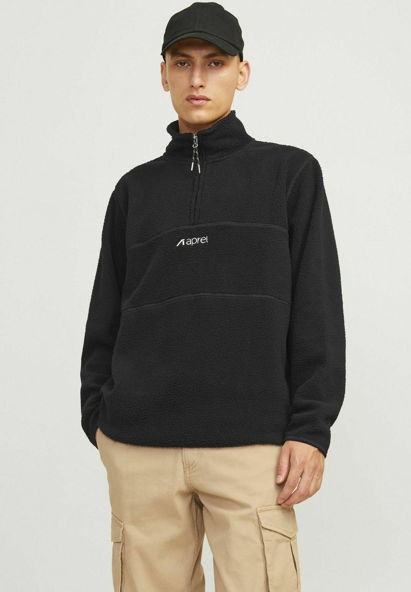 Black fleece quarter-zip pullover features a soft texture, contrasting horizontal seam, and white logo on the chest. Worn with beige cargo pants.