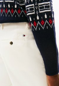Jersey de punto con base azul marino y patrones geométricos en blanco, rojo y negro, combinado con pantalones de pana blancos que tienen un botón y un detalle de logo.