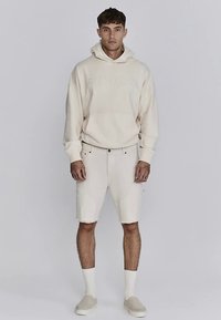 Beige huvtröja med präglad logotyp, kombinerad med ljusbeige jeansshorts. Har en avslappnad passform och minimalistiska detaljer, kompletterad med slip-on skor.