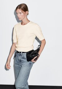 Maglione giallo chiaro a maniche corte accanto a jeans a vita alta blu chiaro. Borsa in pelle nera con design curvato tenuta in mano.