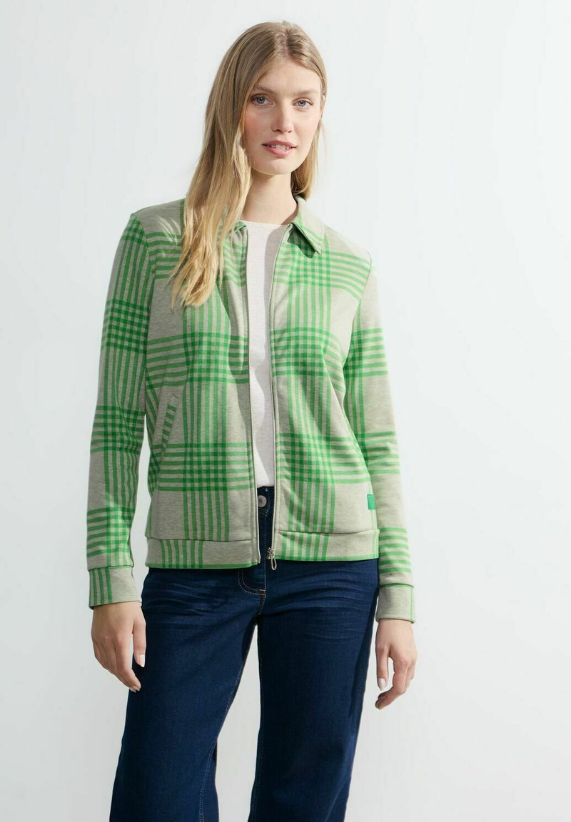 Chaqueta de cuadros verdes con cremallera, que presenta un cuello, mangas largas y puños acanalados, fabricada con un tejido suave. Llevada sobre una blusa blanca y vaqueros azules.