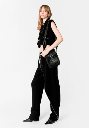 Chaleco negro recortado con cuello alto, combinado con pantalones negros de pierna ancha y zapatos de punta afilada. Pequeño bolso negro de hombro con detalles metálicos.