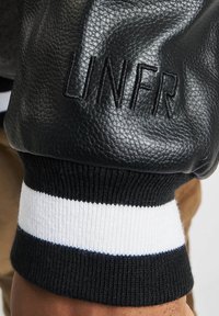 Svart skinn bomberjakke med preget "UNFR" logo. Mansjettene har et svarte og hvite stripete ribbestrikket design. Teksturerte skinnfinish.