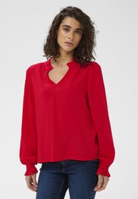 Blouse rouge avec un col en V et un col à volants, manches longues avec poignets froncés, tissu lisse et coupe décontractée.