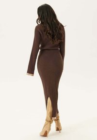 Robe en tricot côtelé brun avec manches longues, forme ajustée, fentes sur les côtés et une fine ceinture. Associée à des bottes à talons hauts beiges.