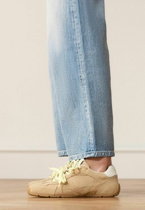 Personne portant un jean bleu clair et des baskets en daim beige, debout sur un sol en bois devant un mur uni.