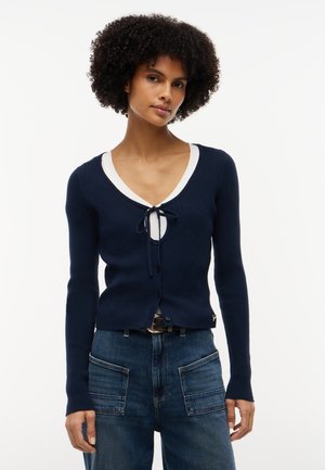 Kvinde med krøllet hår iført en navyblå cardigan med lange ærmer og knapper over en hvid top og blå jeans med store forlommer.
