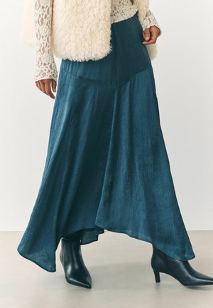 Una gonna maxi fluente di colore teal con una texture increspata, caratterizzata da un orlo alto-basso, abbinata a stivaletti neri con punta a punta.