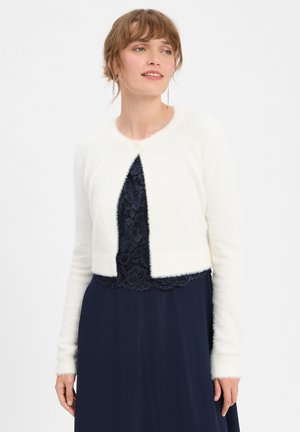 Vrouw draagt een witte, pluizige cropped cardigan over een marineblauwe kanten jurk en kijkt iets opzij tegen een effen witte achtergrond.