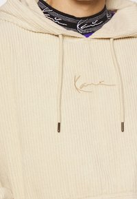 Felpa con cappuccio in corduroy beige, con logo ricamato, coulisse regolabile e texture a costine. Presenta una tasca a marsupio.