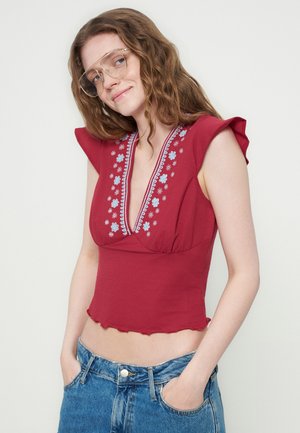 Tânără cu păr ondulat purtând ochelari mari, top roșu brodat fără mâneci cu decolteu adânc în V și blugi albaștri, mâinile în buzunare.