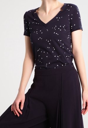 Vrouw draagt een donker V-hals T-shirt met kleine witte driehoekpatronen, gecombineerd met hoge, losse zwarte broek.