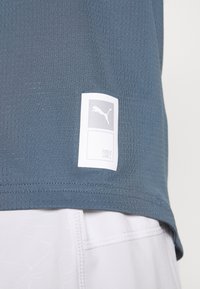 Modrá sportovní triko vyrobené z texturované, prodyšné látky. Obsahuje bílé štítky s logem a textem. Lem je mírně zakřivený.