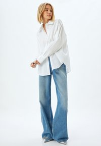 Weißes Langarmshirt mit verzierten Details an den Schultern, kombiniert mit hellblauen weiten Jeans. Silberne Schuhe runden das Outfit ab.