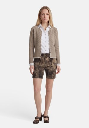 Vrouw die een beige gebreid vest, witte blouse met knopen, traditionele bruine geborduurde leren korte broek en bruine schoenen met hakken draagt, staande naar voren gericht.