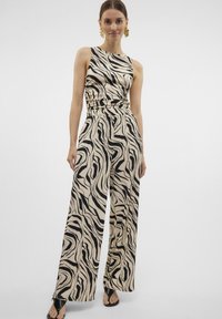 Ärmelloser Jumpsuit in Beige mit schwarzem abstraktem Zebra-Muster, der eine taillierte Taille und einen weiten Beinstil aufweist. Kombiniert mit schwarzen Sandalen.