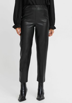 Woman wearing black faux leather slim-fit pants, black long-sleeve blouse with ruffle cuffs, and black pointed-toe ankle boots, standing.