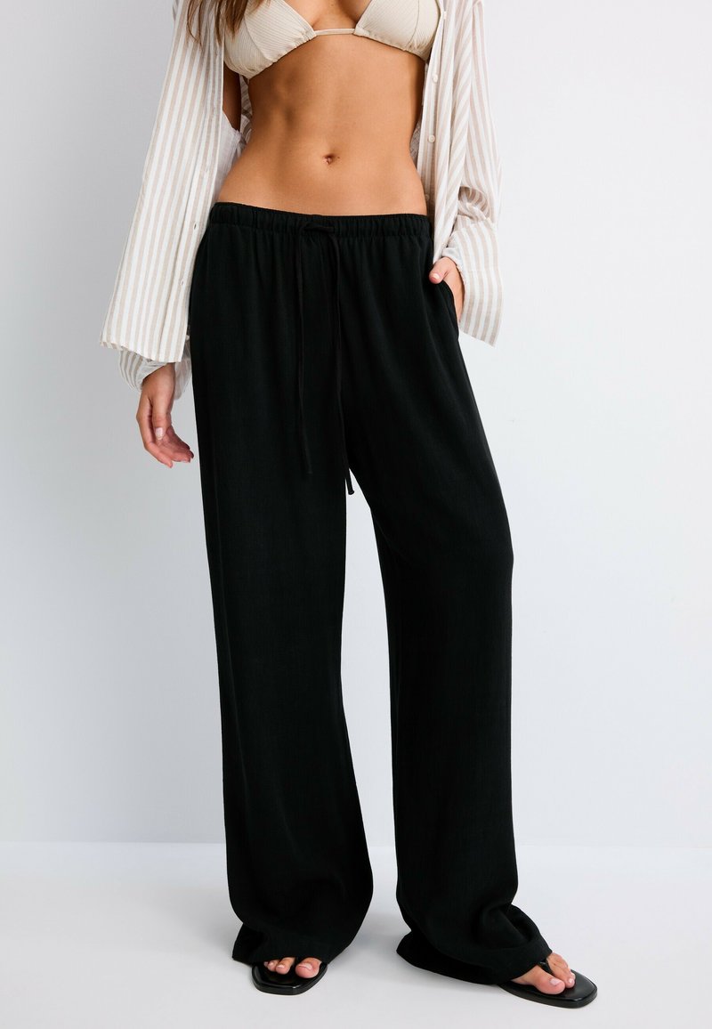 Femme portant un pantalon noir à jambes larges, un haut de bikini beige, une chemise rayée ouverte et des sandales noires à bout carré sur un fond clair uni.