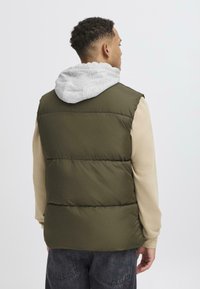 Maslinasto zeleni puffer prsluk s sivim kapuljačom, stražnji pogled prikazuje kviltanu teksturu i bež majicu dugih rukava ispod, ležeran stil.