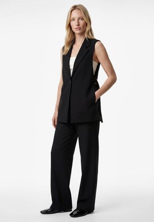 Mujer de pie, con un blazer negro sin mangas sobre una blusa blanca, pantalones negros de pierna ancha y zapatos negros puntiagudos, posando con una mano en el bolsillo.