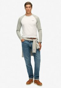 Biała koszula z długim rękawem i szarymi rękawami raglanowymi, niebieskie dżinsy i brązowe buty. Szary sweter związany w pasie dodaje charakteru.