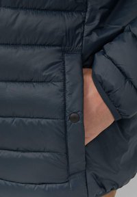 Marineblaue isolierte Jacke mit einem gesteppten Design, Seitentasche mit einem schwarzen Knopf und gerippten Bündchen. Glatte, glänzende Textur.