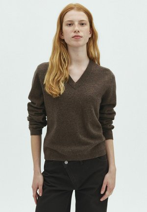 Jeune femme aux longs cheveux rouges portant un pull marron à col en V et un pantalon noir, debout devant un fond clair uni.