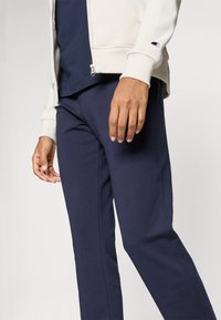 Lys beige zip-up hettegenser med ribbestrikkede mansjetter, kombinert med marineblå joggebukser. Begge laget av mykt stoff, med en logo på ermene.