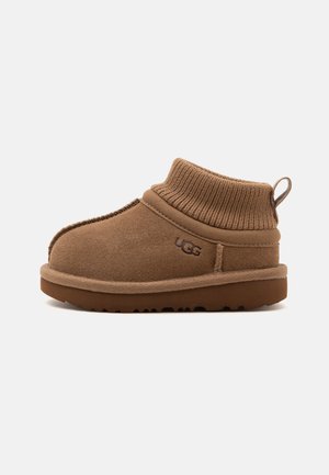 Bottine en daim marron à la cheville avec un bord-côte côtelé, languette à l’arrière et logo UGG sur le côté, présentée de profil sur fond blanc.