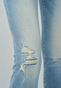 Jeans de color azul claro con efecto desgastado, que presentan un desgarro en la rodilla con bordes deshilachados y franjas tejidas visibles en el interior de la rotura. Textura de mezclilla suave.