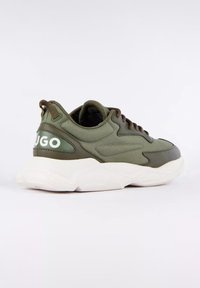 Baskets basses vert olive avec une semelle blanche épaisse et des lacets marron, comportant la marque "HUGO" au niveau du talon.