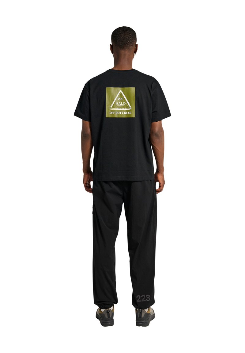 Mann trägt ein schwarzes T-Shirt mit grünem Dreieck-Logo und dem Schriftzug "HALO 223 OFF DUTY GEAR" auf dem Rücken, kombiniert mit schwarzer Hose und Turnschuhen.