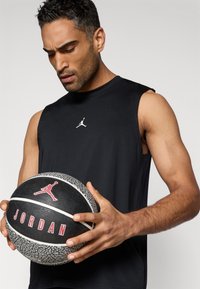 Μαύρο αθλητικό τοπ χωρίς μανίκια με λευκό λογότυπο Jumpman. Το άτομο κρατάει μια υφασμάτινη μαύρη μπάλα μπάσκετ με κόκκινα γράμματα "JORDAN" και λογότυπο.