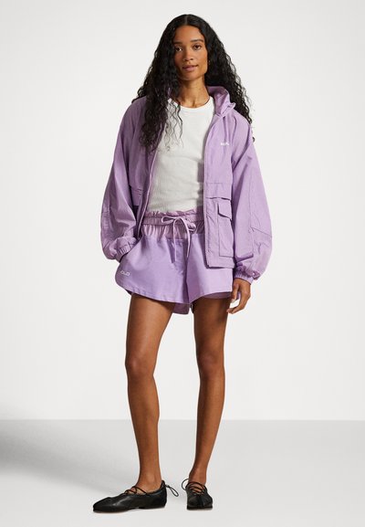 Γυναίκα με μακριά σγουρά μαλλιά, που φοράει λευκό τοπ, λιλά φαρδιά μπουφάν και ταιριαστά σορτς, καθώς και μαύρα δερμάτινα παπούτσια με κορδόνια, στέκεται μπροστά σε λευκό φόντο.