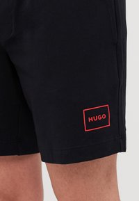 Shorts negri din bumbac, cu o textură netedă. Prezintă un logo brodat roșu, etichetat "HUGO", pe partea inferioară a piciorului stâng.